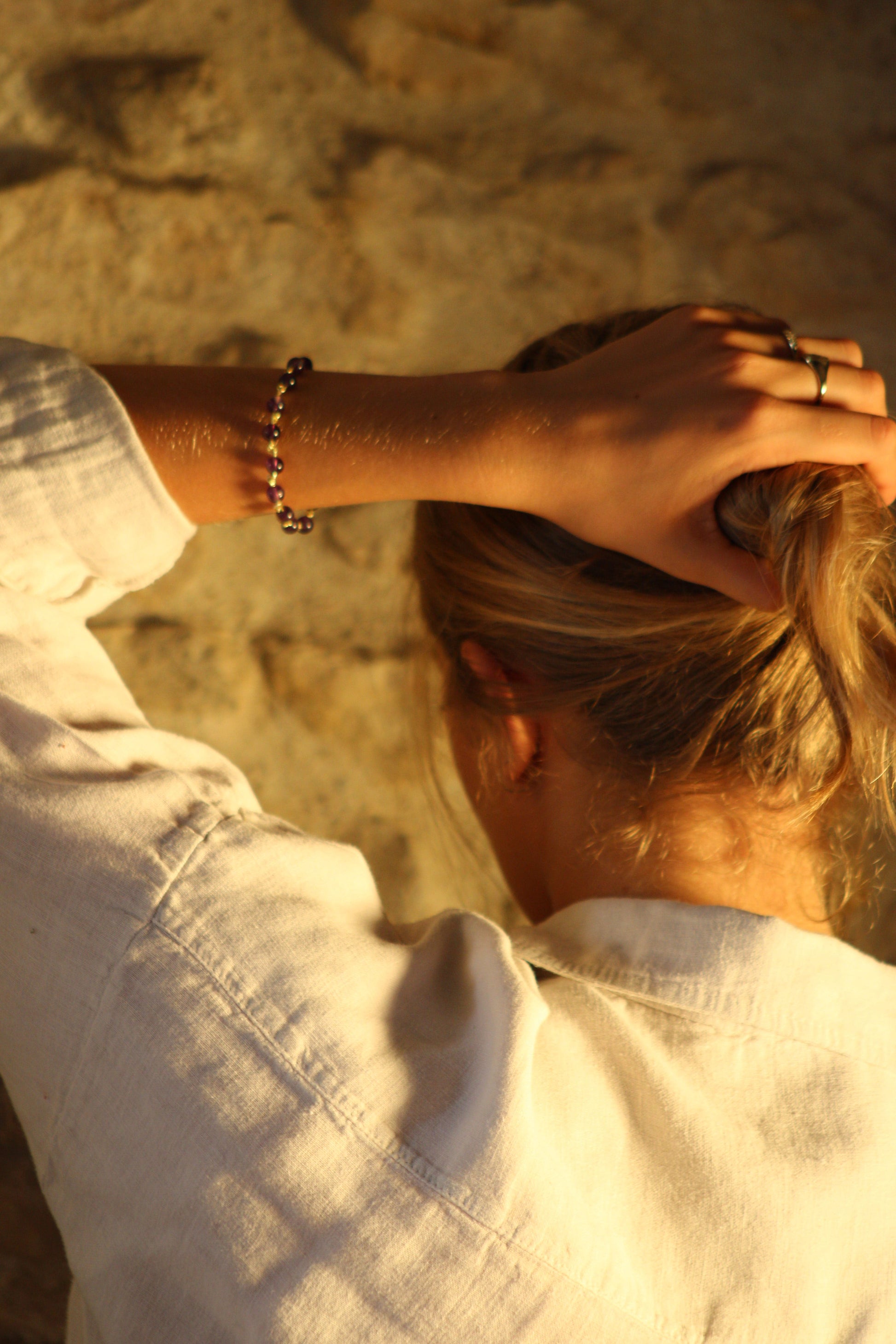 photo d'une femme tenant ses cheveux sur un fond de mur en pierre avec un bracelet Ornéa en pierres naturelles d'améthyste au poignet